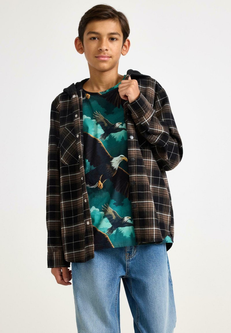 Svart- och brunt rutig flanellskjorta över en turkos grafisk t-shirt med örnar, ihop med ljusblå jeans. Avslappnad lager-på-lager outfit.