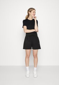 T-shirt in cotone nero abbinato a pantaloni da ginnastica neri di lunghezza media, fino alla coscia. I pantaloni presentano una vita elastica con cordino e un accento con logo. Sneakers bianche.