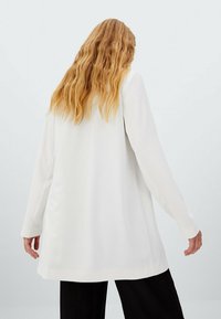 Personne aux cheveux longs et ondulés portant un long blazer blanc et un pantalon noir, vue de dos sur un fond uni.