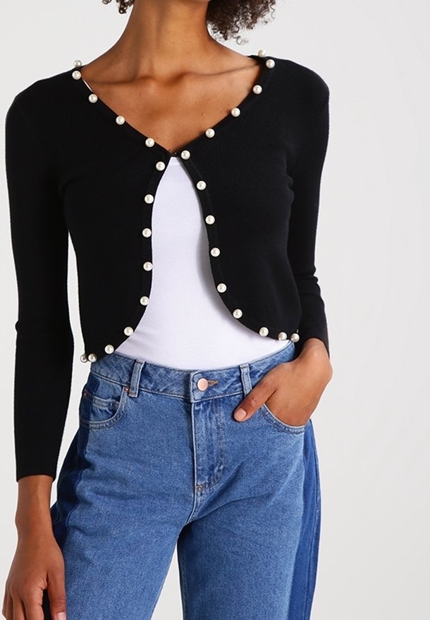 Femme portant un cardigan noir court avec des boutons en perle sur un haut blanc et un jean bleu taille haute, main dans la poche.