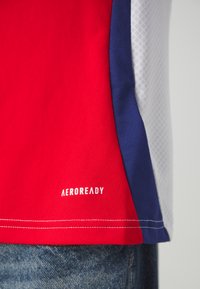 Červené sportovní tričko s modrými a bílými akcenty. Má síťovou strukturu a logo "AEROREADY" blízko lemu, detaily dvojitého prošití.