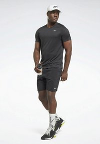 Svart treningsskjorte med korte ermer og svarte shorts, brukt sammen med hvit caps og svarte joggesko med neonfargede detaljer, holder en vannflaske.