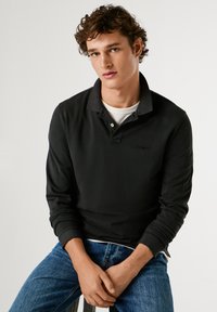 Černé polokošile s dlouhým rukávem a límečkem, lehký materiál a detail loga. V kombinaci s modrými džínami, v posedě.