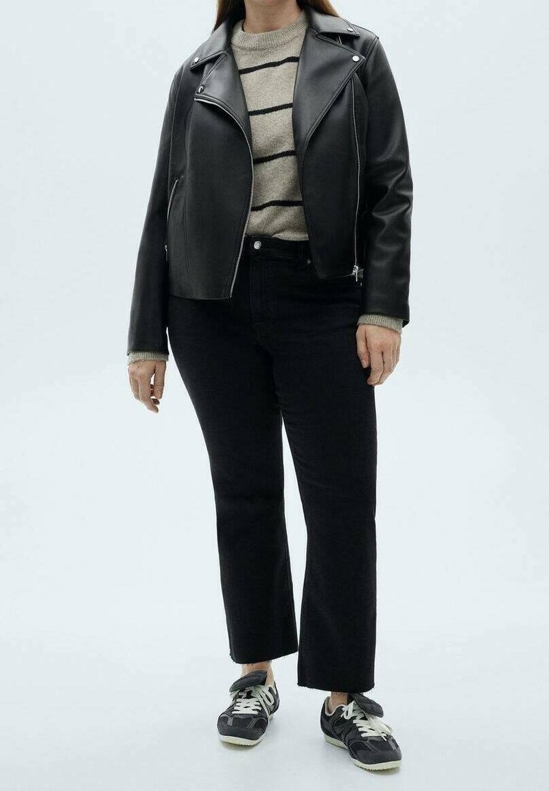 Personne portant une veste en cuir noire, un pull beige à rayures, un pantalon court noir et des baskets noires et blanches, debout sur un fond uni.