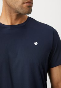 Κοντινή λήψη ενός άνδρα που φοράει ένα ναυτικό μπλε υφασμάτινο T-shirt με ένα μικρό λευκό κυκλικό λογότυπο στο αριστερό μέρος του στήθους.