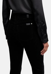 Jeans en velours noir avec une coupe ajustée, dotés de poches arrière et d'une étiquette de marque discrète. Assortis à une chemise noire en satin à manches longues.