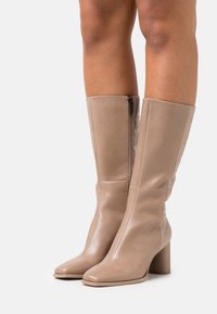 Botas de cuero marrón hasta la rodilla con tacones gruesos, usadas sobre piernas bronceadas, con cremalleras laterales y una textura suave.