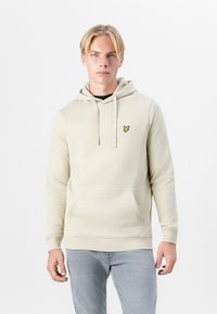 Hoodie beige avec cordon de serrage, poche kangourou et écusson jaune. Fabriqué en tissu doux ; présente des poignets et un ourlet côtelés.