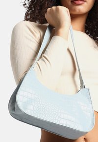 Ljusblå handväska i konstläder med krokodilmönstrad textur, böjd form och en lång rem, hålls av en person i en krämfärgad topp.