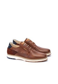 Zapatos de cuero marrón con textura perforada, tacón oscuro contrastante, suela blanca y diseño de cordones. Acento de marca notable a un lado.
