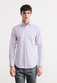 Νεαρός άνδρας που φοράει ανοιχτό μωβ πουκάμισο με μακριά μανίκια και γκρι παντελόνι, στέκεται μπροστά σε ένα απλό λευκό φόντο.