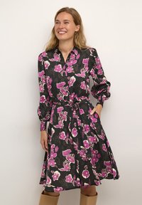 Frau lächelt in einem schwarzen Blumenkleid mit rosa und weißen Blumen, langen Ärmeln, einem Taillengürtel und kniehohen, sandfarbenen Stiefeln.