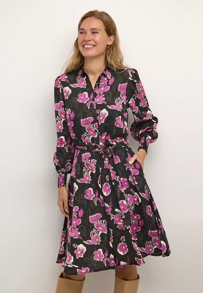 Frau lächelt in einem schwarzen Blumenkleid mit rosa und weißen Blumen, langen Ärmeln, einem Taillengürtel und kniehohen, sandfarbenen Stiefeln.
