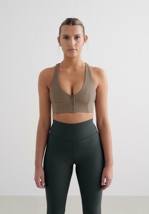 Beha in lichtbruin, geribbelde stof met een voering; gecombineerd met hoge donkergroene leggings. Soepele textuur, atletisch ontwerp.