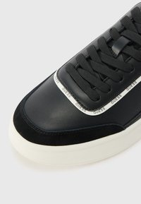 Sort sneakers med glat læder- og ruskindmateriale, flade sorte snørebånd, hvid mellemsål og en logo-detalje på siden.