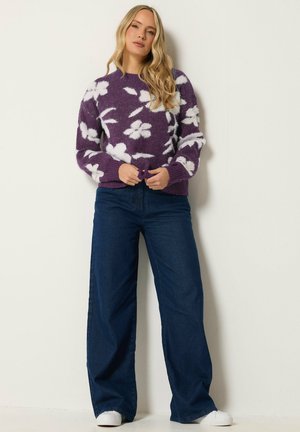 Frau mit langen blonden Haaren, die einen lila Pullover mit weißen Blumenmustern, weite dunkelblaue Jeans und weiße Schuhe trägt, steht an einer weißen Wand.
