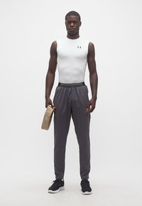 Άνδρας με λευκό μπλουζάκι συμπίεσης χωρίς μανίκια και γκρι αθλητικό παντελόνι κρατάει ένα καφέ λαστιχένιο ελατήριο, στεκόμενος μπροστά σε απλό λευκό φόντο.