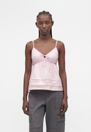 Vrouw met gevlochten haar die een lichtroze mouwloos top met strikdetail en grijze cargobroek draagt, staand voor een effen witte achtergrond.