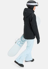 Snowboarder in black jacket, light blue pants, and black boots, holding a white snowboard with blue lettering, wearing a black helmet and goggles.