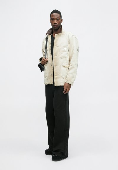 Veste beige à boutons avec un col marron, dotée de deux poches poitrine et d'une finition texturée, associée à un pantalon noir à larges jambes et des chaussures.