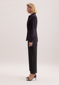 Kobieta o blond włosach w dopasowanej czarnej bluzce z długim rękawem, czarnych spodniach o prostych nogawkach oraz czarnych wysokich butach z odkrytymi palcami, stojąca bokiem.