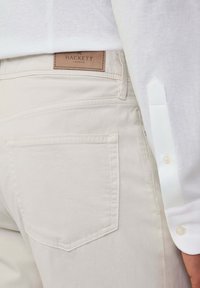 Beige katoenen broek met een leren label met de tekst "Hackett London." Voorzien van achterzakken en witte stiksels. Draagt een wit overhemdbloes.