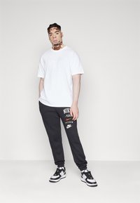 Biele Nike tričko, šedé Nike tepláky s logom a čierno-biele Nike tenisky. Model stojí na plain pozadí.