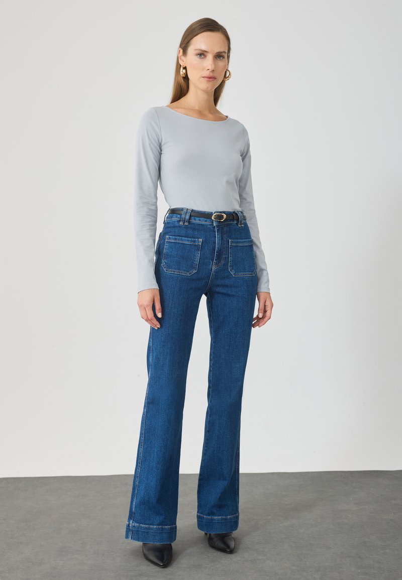 Maglietta a maniche lunghe grigia chiara abbinata a jeans a vita alta blu scuro a campana, con tasche anteriori e una cintura nera con fibbia argentata.