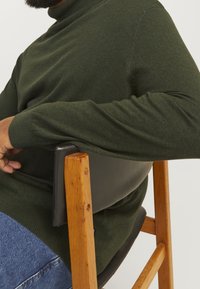 Zielony sweter z golfem wykonany z miękkiego materiału, z fakturowaną powierzchnią i dopasowanymi ściągaczami, noszony podczas siedzenia na minimalistycznym drewnianym krześle.