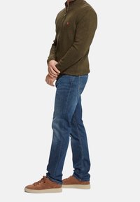 Uomo che indossa un maglione verde oliva con zip, jeans blu e scarpe da ginnastica marroni, in piedi con le mani giunte davanti su uno sfondo bianco.