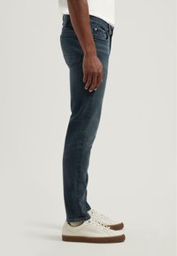 Slim-fit tmavě modré džínové kalhoty s hladkou texturou, středním pasem, pěti kapsami a mírně vybledlým vzhledem. V kombinaci s bílými teniskami.