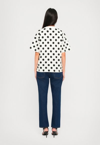 White short-sleeve top with black polka dots, paired with blue straight-leg jeans and black heeled sandals, viewed from the back.