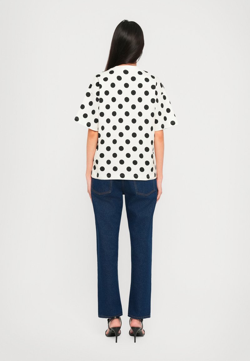 Witte top met korte mouwen en zwarte stippen, gecombineerd met blauwe straight-leg jeans en zwarte hoge hakken, gezien van achteren.
