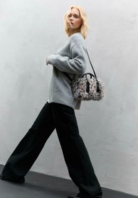 Donna bionda che cammina contro un muro grigio, indossando un sweater grigio, pantaloni neri a gamba larga e portando una piccola borsa in finta pelliccia maculata con manici neri.