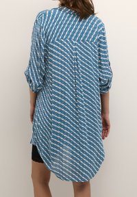 Chemise bleu à manches longues avec un motif blanc, boutonnée à l'arrière et à ourlet arrondi. Fabriquée en tissu léger, elle présente des manches retroussées.