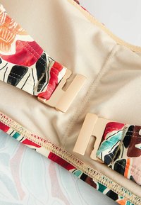 Béžové plavky s farebnými kvetinovými ramienkami a zlatými sponami ležia položené na vzorovanej ploche.