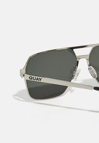 Pari aviator aurinkolasit, joissa on metallinhohtoinen hopeanvärinen runko, tumma vihreä linssi ja kirkkaat nenätyynyt; rungossa on "QUAY"-logo.