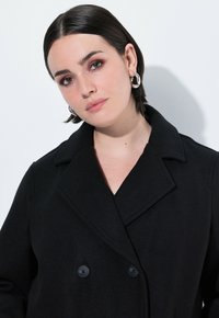 Černý dvouřadý kabát s širokým límcem a texturovanou tkaninou. Obsahuje kulaté stříbrné náušnice a neutrální pozadí.