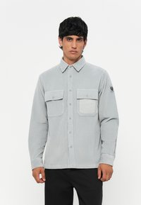 Light gray corduroy shirt with long sleeves, buttoned front, two chest pockets, and logo patch on the left sleeve. Smooth, textured fabric.