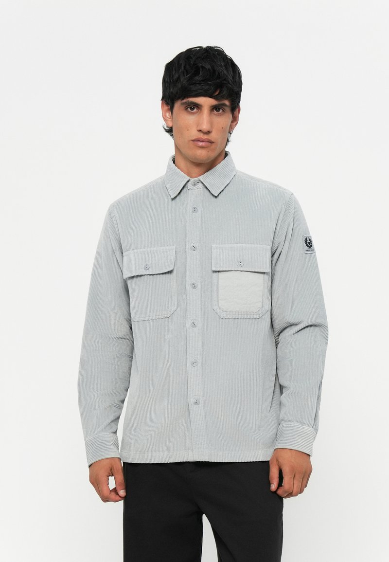 Light gray corduroy shirt with long sleeves, buttoned front, two chest pockets, and logo patch on the left sleeve. Smooth, textured fabric.