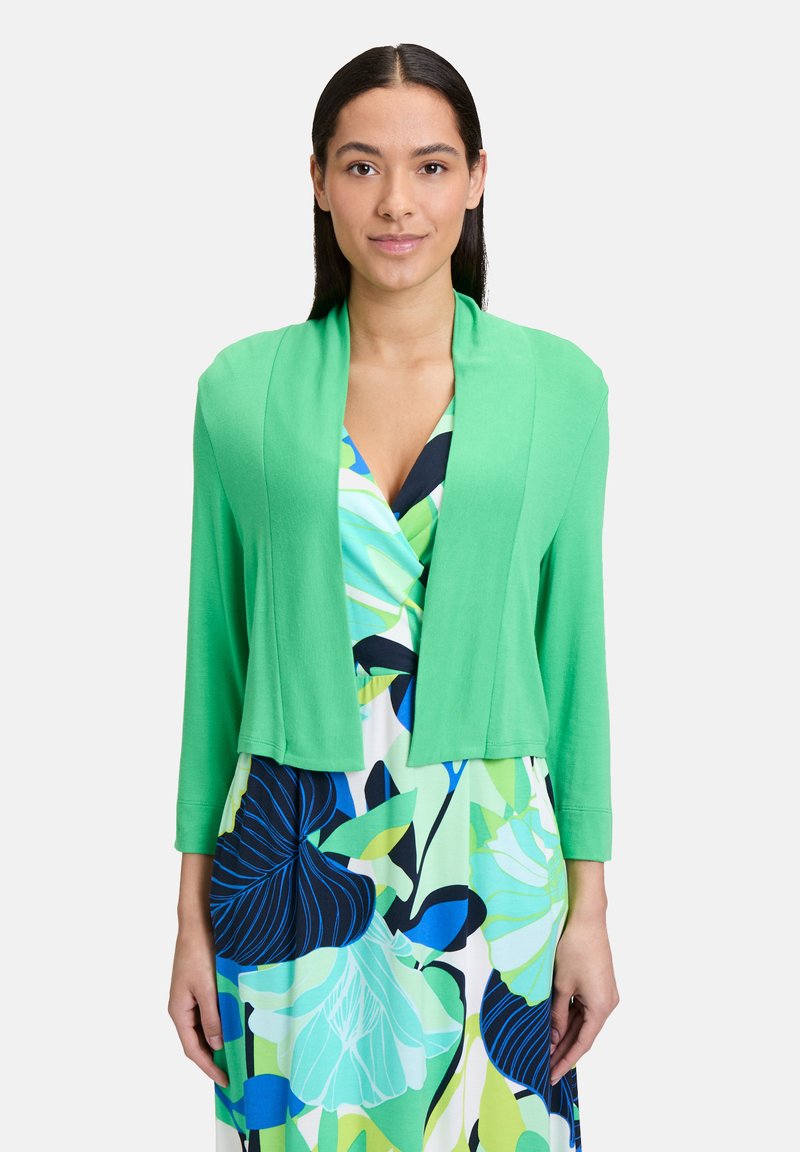 Cardigan vert court avec une texture douce et un design à front ouvert. Des manches longues complètent une robe florale multicolore en dessous.