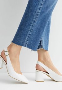 Osoba oblečená v modrých džínách a bílých sandálech na blokovém podpatku se zlatou přezkou, stojící na bílé ploše.