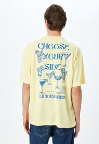 Camiseta amarilla de manga corta con gráficos azules de varios cócteles y el texto "ELIGE TU LADO" en la parte posterior. Material texturizado.