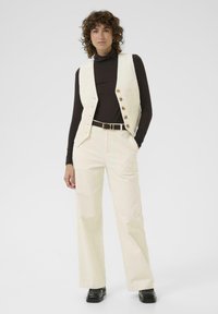 Crème corduroy gilet met vijf knopen, gecombineerd met bijpassende wijde broek. Zwarte coltrui eronder en zwarte schoenen.