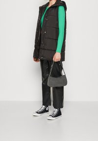 Schwarze Steppweste mit Kapuze über einem grünen gerippten Langarmoberteil, kombiniert mit schwarzen Lederhosen und schwarzen Sneakers. Graue Handtasche.