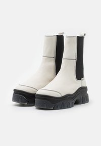 Botines gruesos con una parte superior de cuero blanco, paneles laterales elásticos negros y una suela de goma negra texturizada. Costuras de contraste visibles.