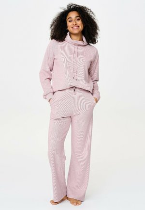 Woman with curly hair stands barefoot wearing a soft pink long-sleeve top with drawstrings and matching wide-leg pants with pockets.