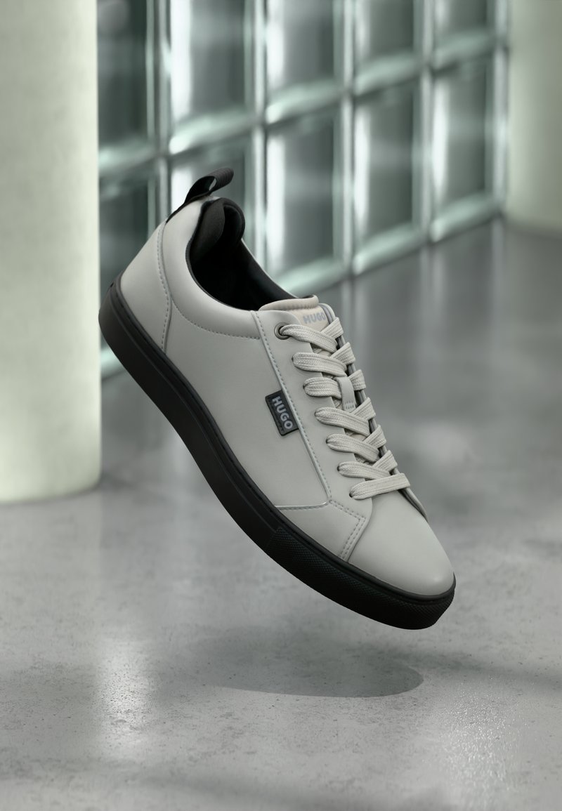 Baskets gris clair en cuir lisse, avec des accents noirs et une semelle en caoutchouc. Les lacets sont soigneusement noués et une étiquette de la marque est visible.