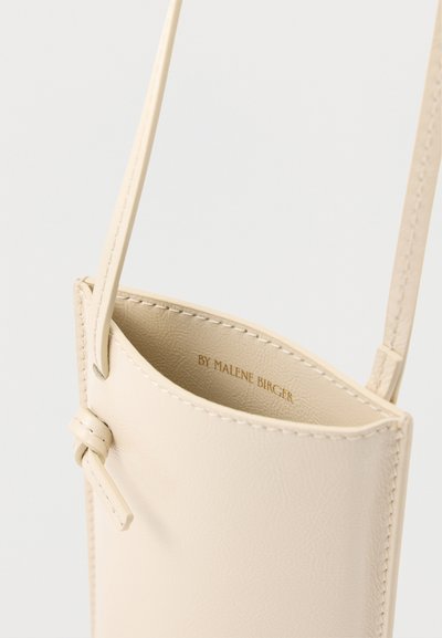 Μικρή μπεζ δερμάτινη τσάντα χιαστί με εμφανείς ραφές και λεπτομέρεια δέσιμου λουριού, κρεμασμένη σε ανοιχτό φόντο.