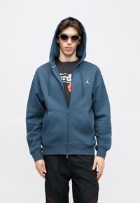 Sweat à capuche zippé en tissu bleu foncé, avec une poche avant, des poignets côtelés et un petit logo blanc sur la poitrine gauche.